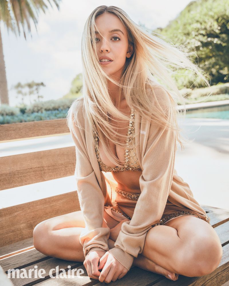 Candid outdoor portrait of blonde woman sitting cross-legged on a wooden bench wearing a gold bikini top, sweater, and skirt.