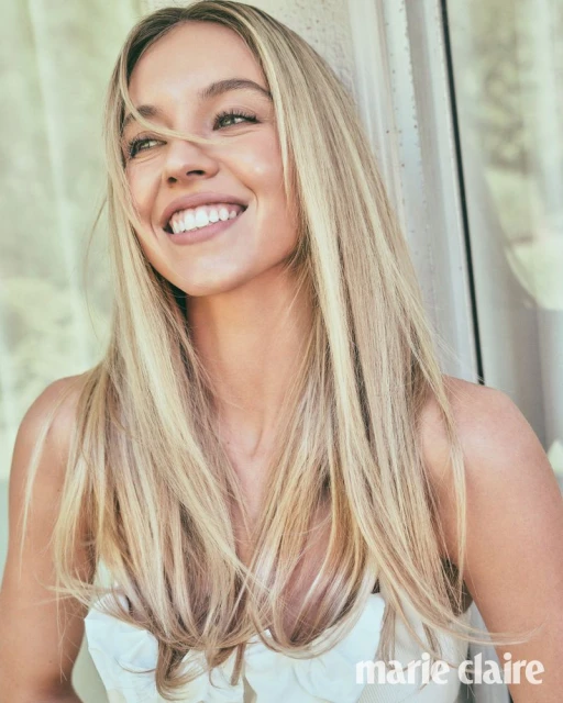 Smiling Sydney Sweeney, a fair-skinned blonde woman, poses in an eye-level medium shot with a window frame visible in the background.