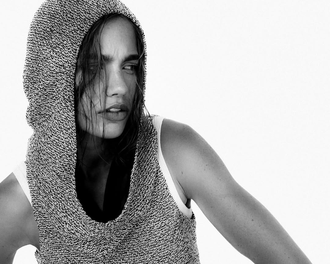 Black and white portrait of an adult woman in a knitted hood and tank top against a bright white background looking off to the side.
