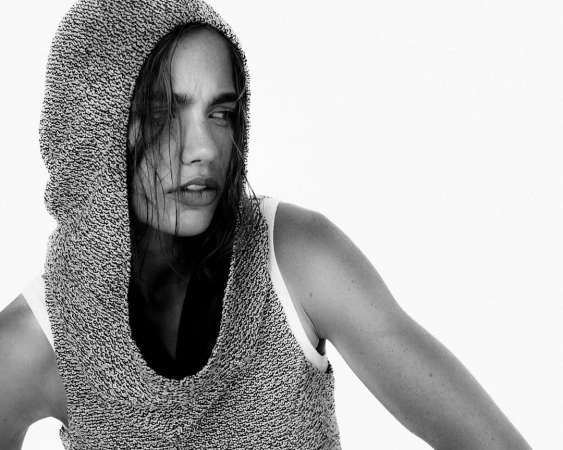 Black and white portrait of an adult woman in a knitted hood and tank top against a bright white background looking off to the side.