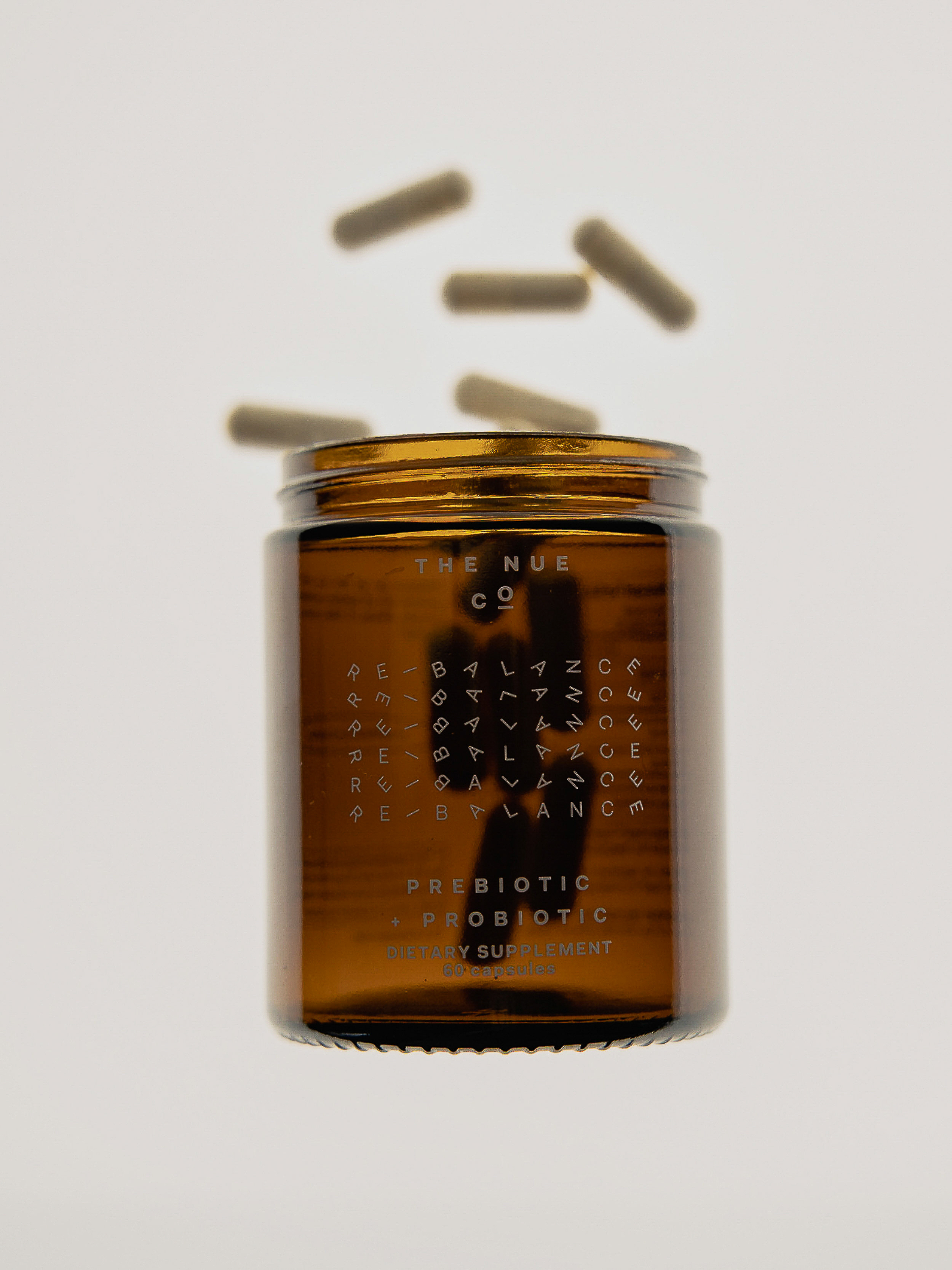 Amber glass jar of prebiotics by The Nue Co. with capsules against a white backdrop. Focus on product, clean and minimalist design.