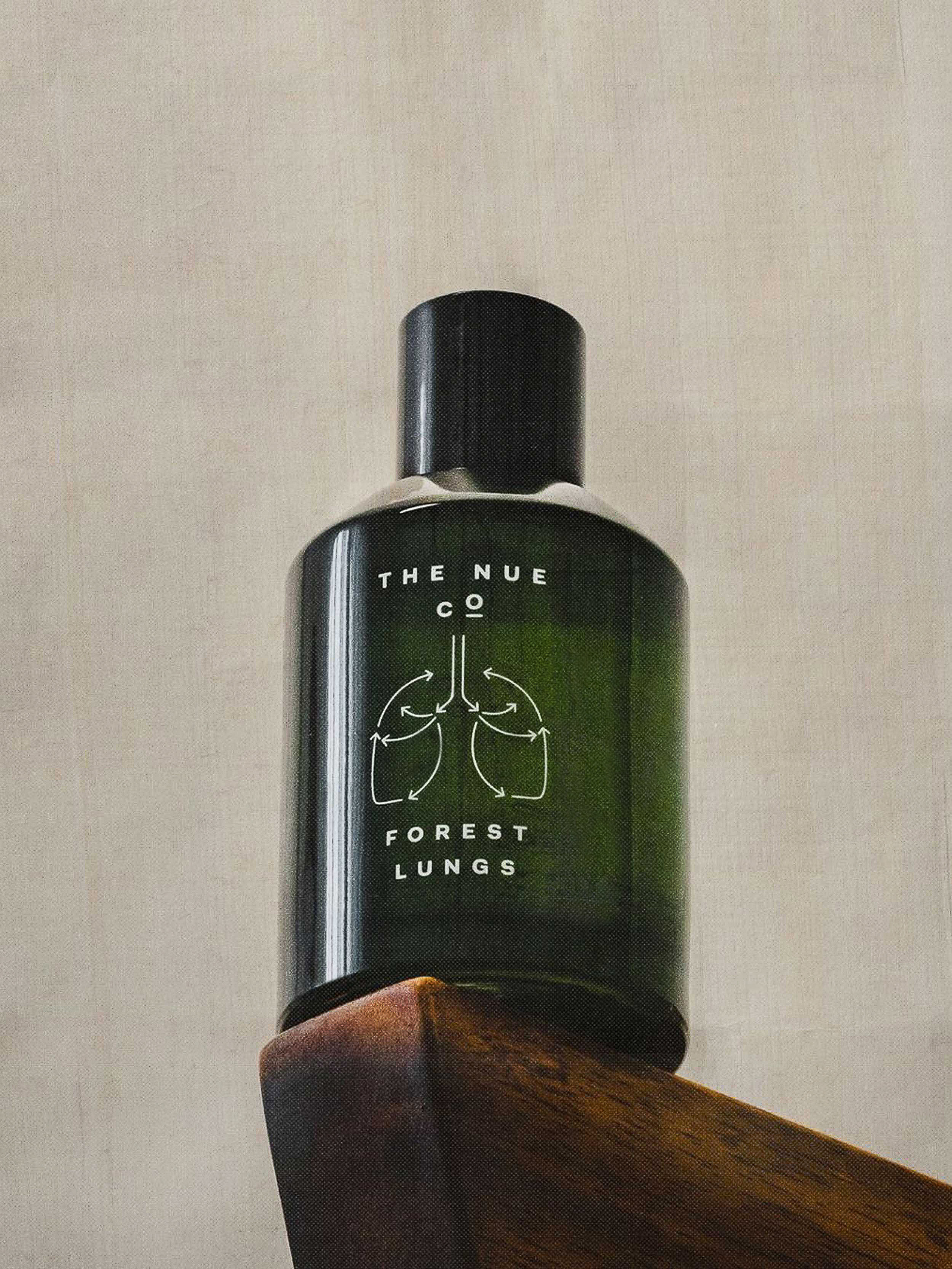 Close-up shot of a green glass bottle of 'Forest Lungs' by The Nue Co. set on a dark wood chair against a neutral background.