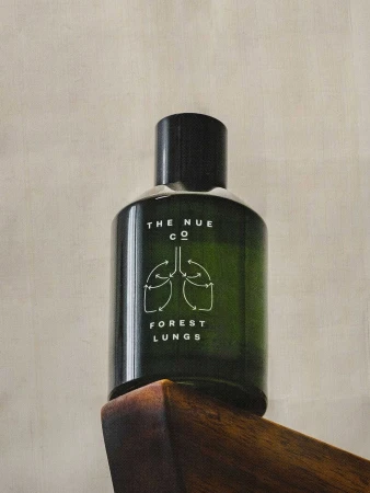 Close-up shot of a green glass bottle of 'Forest Lungs' by The Nue Co. set on a dark wood chair against a neutral background.