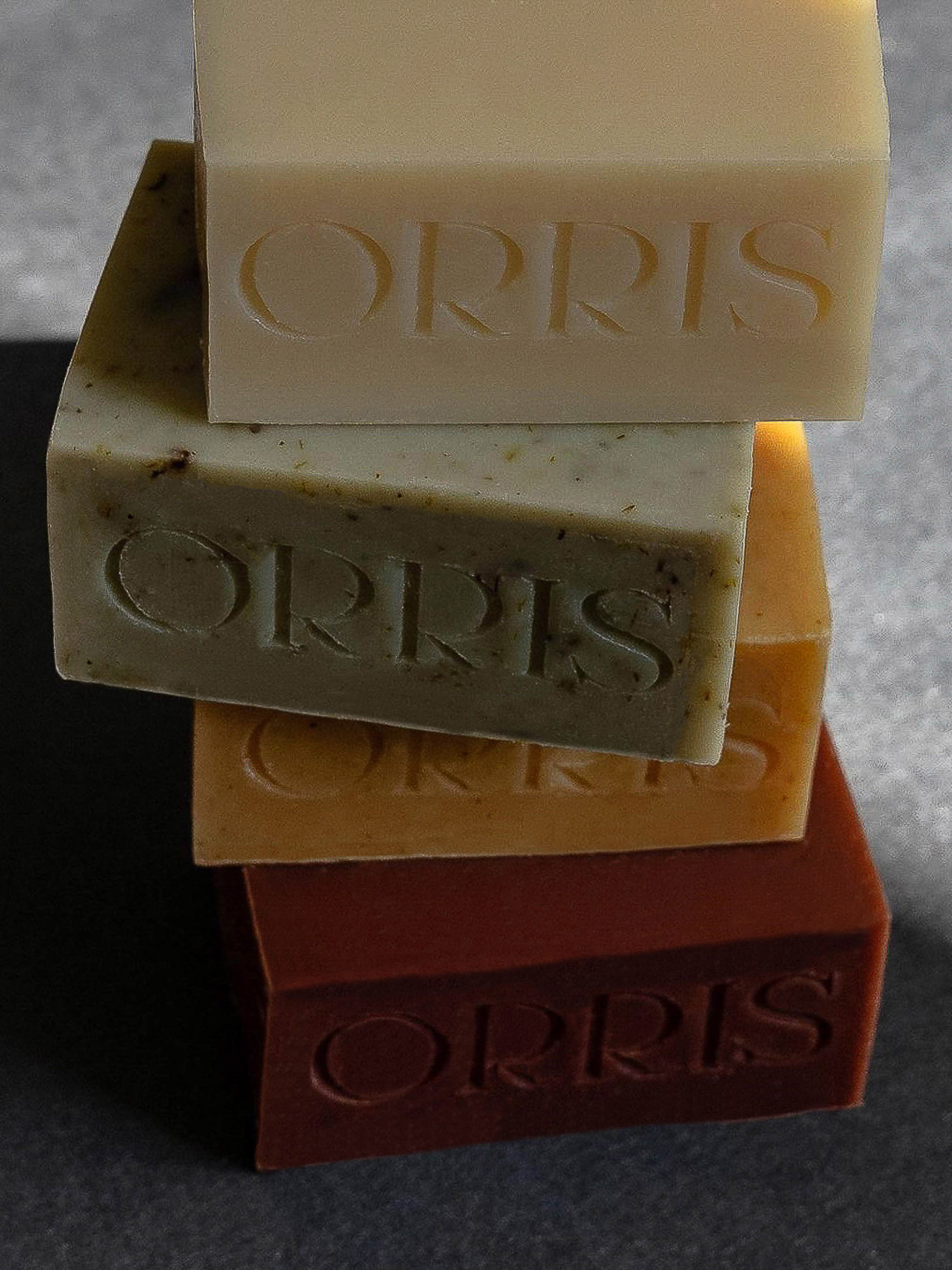Stack of four Orris brand soap bars in different colors on a gray surface in soft lighting, great for beauty or personal care ad.