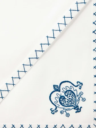 Close-up of an ornate, white fabric napkin with decorative cross-stitch edging and a stylized, floral-paisley design embroidered in dark blue thread.