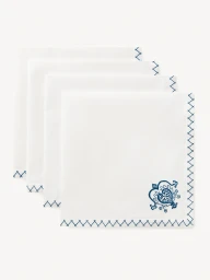 Four folded white cloth napkins with blue embroidered cross-stitch borders stacked against a plain light studio background.