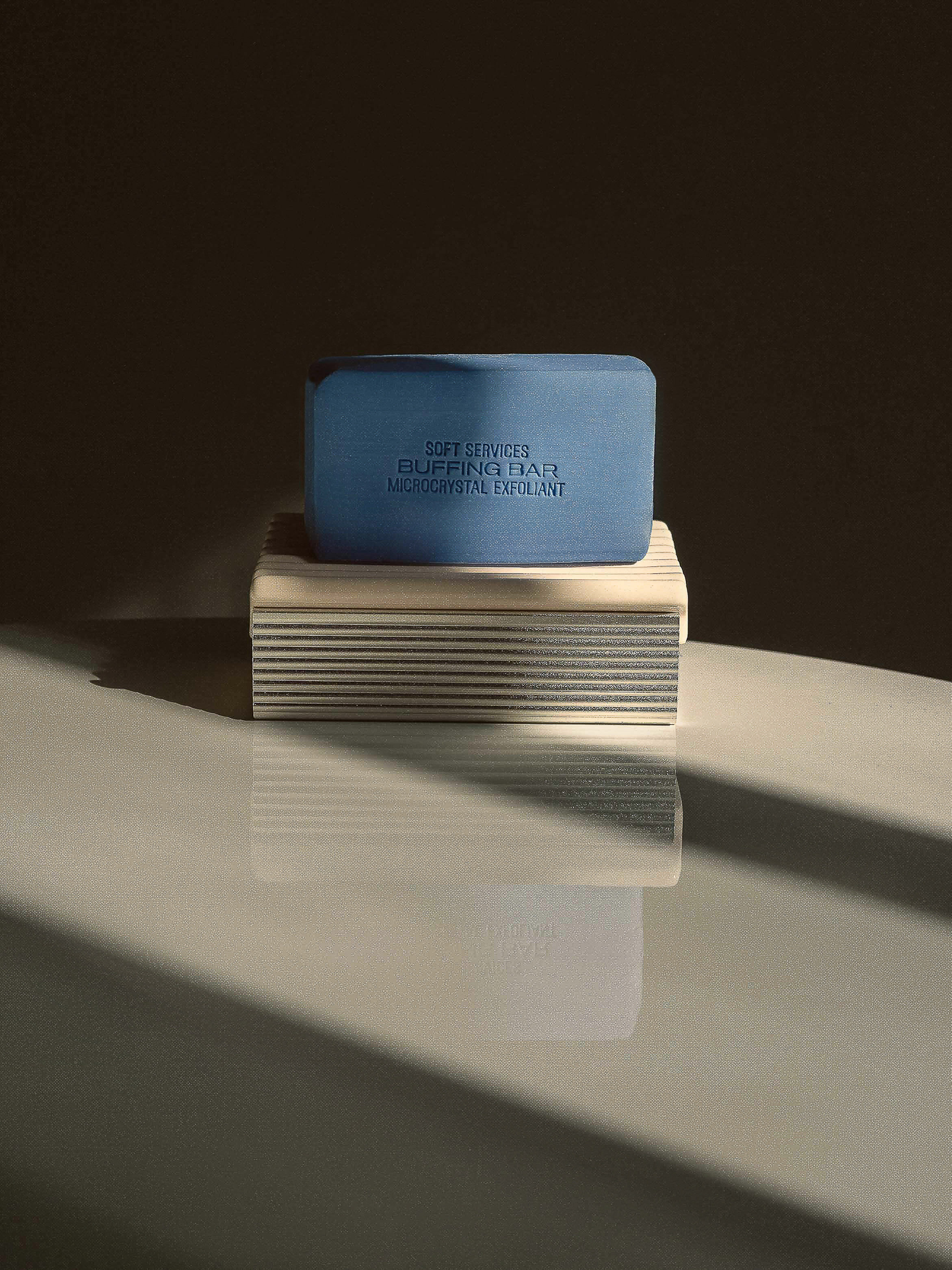 Blue bar soap sits on a textured platform, reflecting in the glossy surface below, captured in soft lighting with dark shadows.