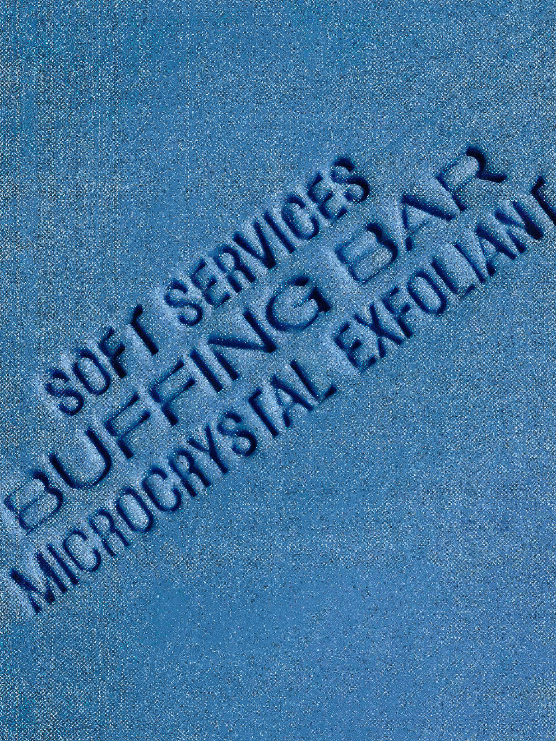 Text on a blue surface reads: SOFT SERVICES, BUFFING BAR, MICROCRYSTAL EXFOLIANT in a slightly angled orientation, creating a skincare product advert.