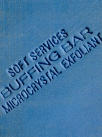 Text on a blue surface reads: SOFT SERVICES, BUFFING BAR, MICROCRYSTAL EXFOLIANT in a slightly angled orientation, creating a skincare product advert.