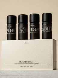 Four black Vitruvi essential oil bottles standing behind a textured cream Signature Kit box on a warm neutral surface.
