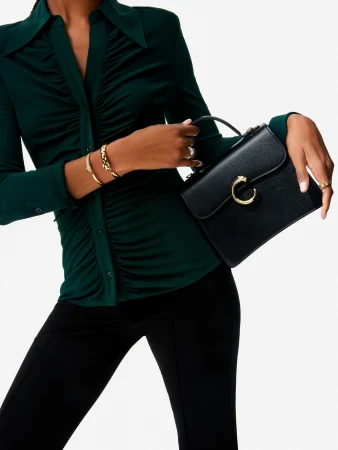 A woman in a dark green ruched shirt holds a structured black leather handbag with a gold clasp against a white seamless background.