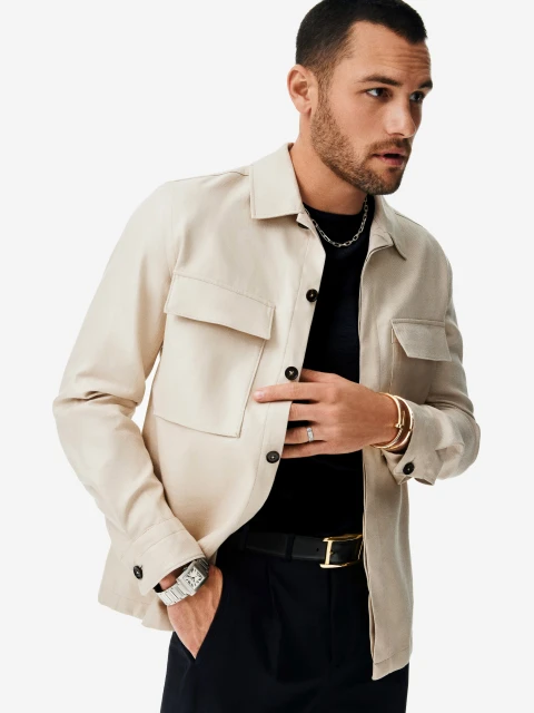 Stylish adult male wearing a beige jacket and black pants, accessorized with a silver chain, bracelets, and a watch, posing indoors on white.
