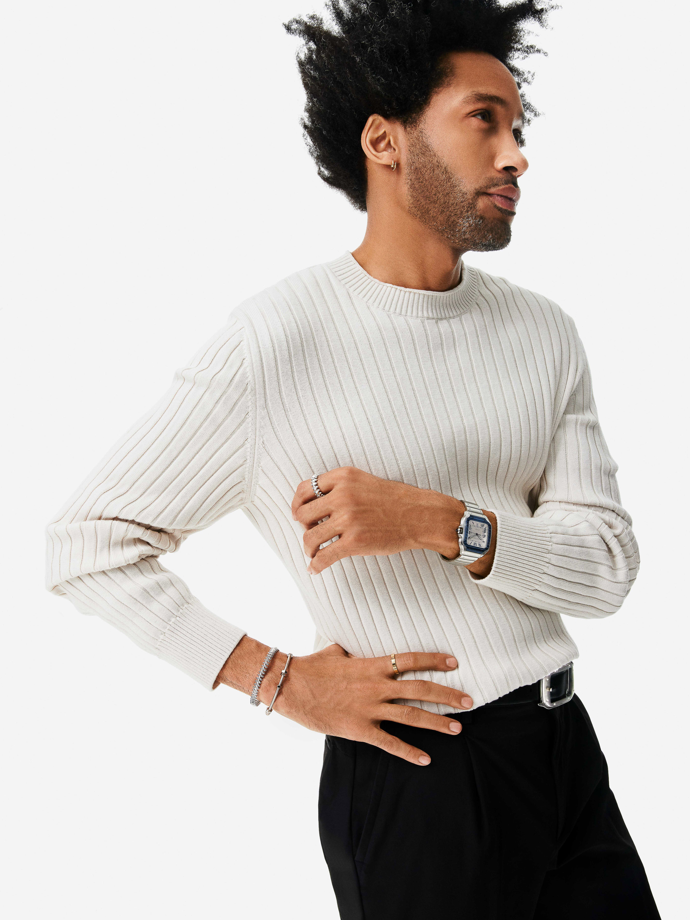 Black man wearing a cream ribbed knit sweater and silver luxury watch against a white seamless studio backdrop.