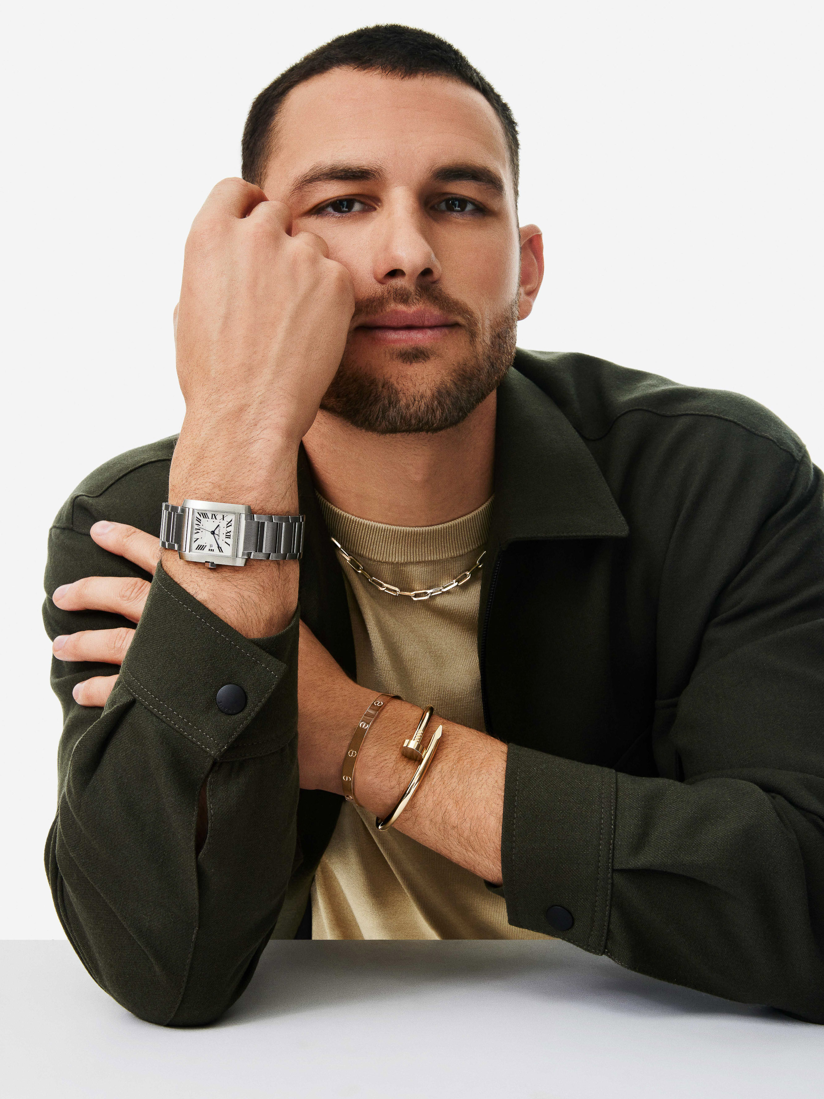 Adult male poses in a photo studio wearing a dark green collared shirt, gold chain and bracelet, and a stainless steel wristwatch.