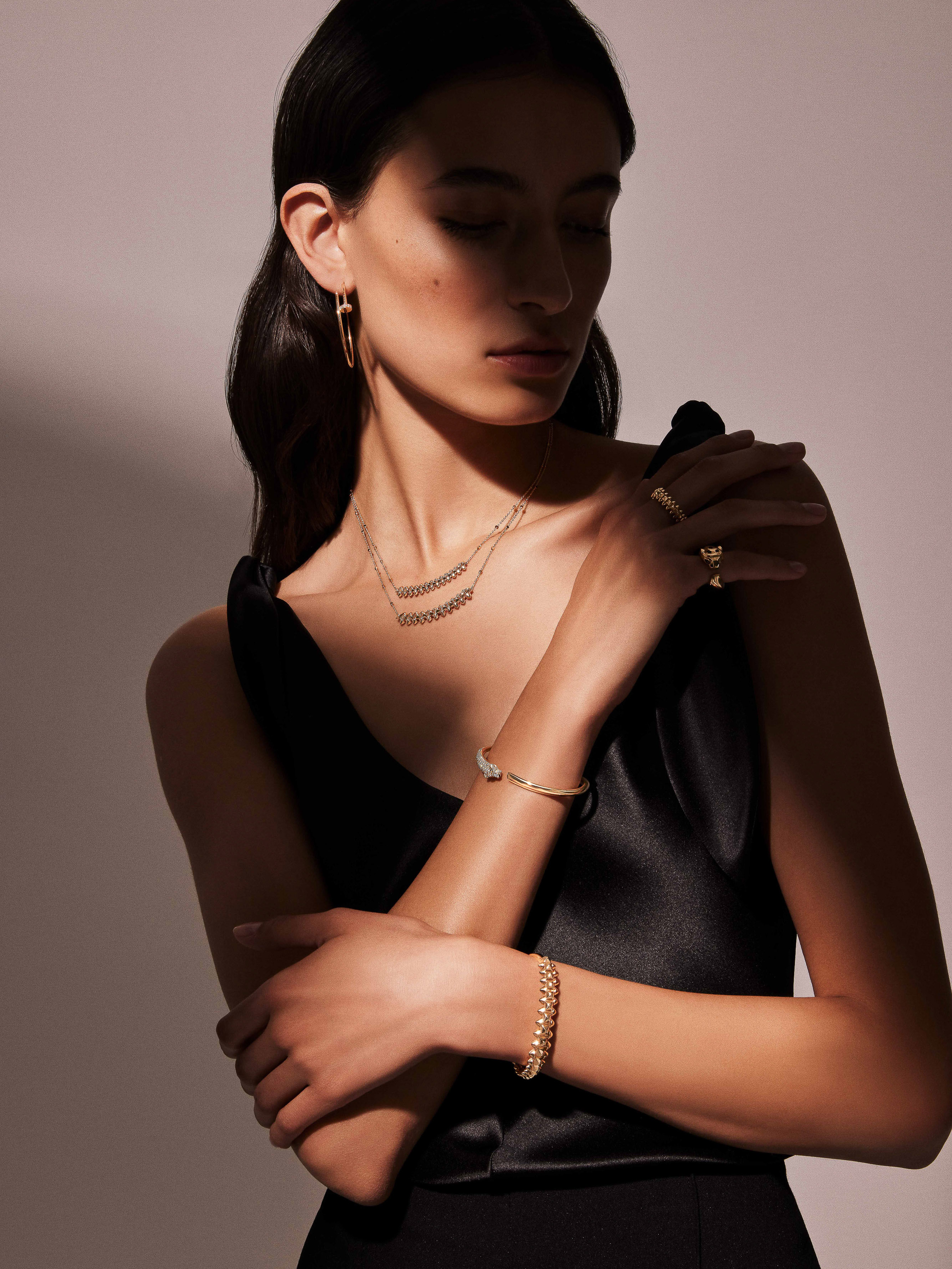 Woman in a black dress poses, showcasing gold jewelry: earrings, necklace, and bracelets. Studio shot, soft lighting. Fashion, beauty concept.