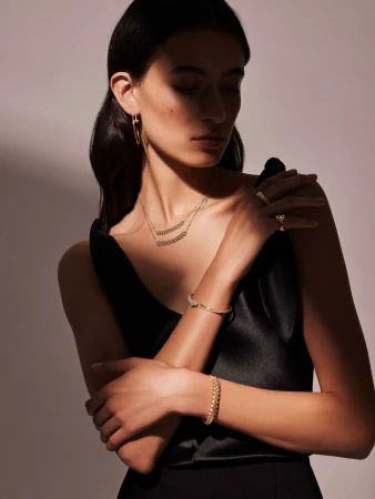 Woman in a black dress poses, showcasing gold jewelry: earrings, necklace, and bracelets. Studio shot, soft lighting. Fashion, beauty concept.