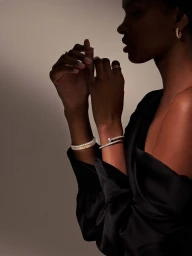 Black woman in an off-the-shoulder black satin top showcasing diamond bracelets and rings in warm low-key lighting.