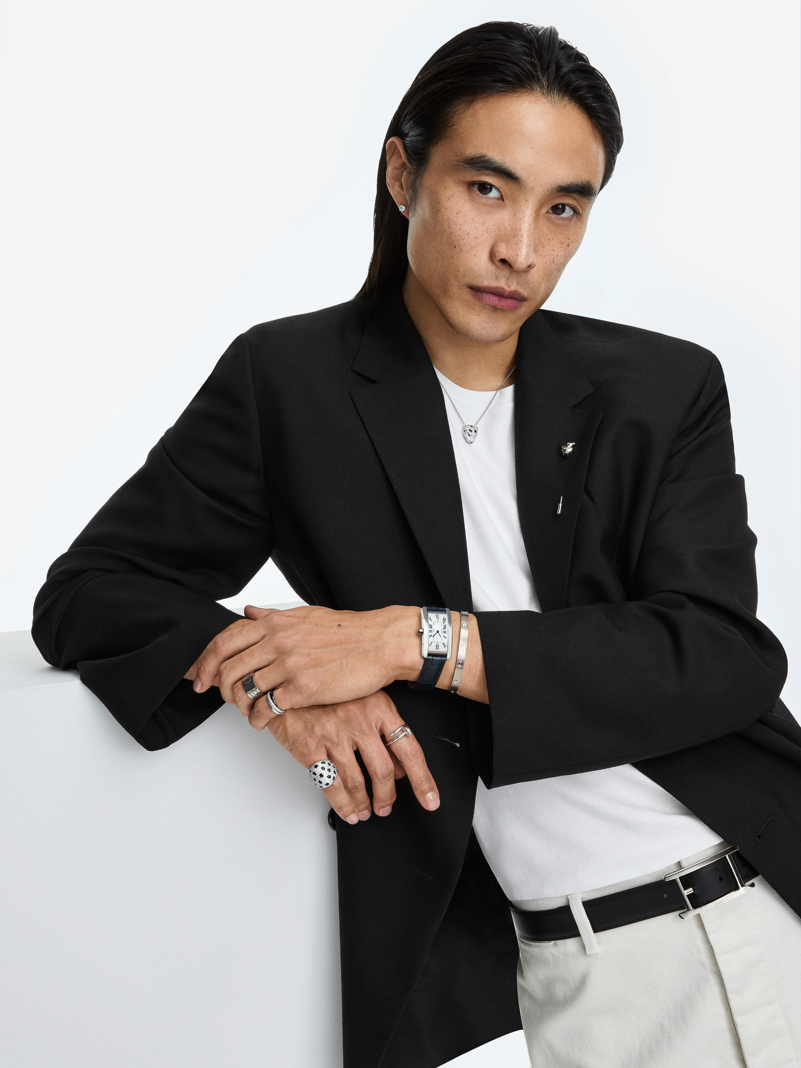 Portrait of a male model with long dark hair wearing jewelry, a black jacket, and white pants in a brightly lit indoor studio.