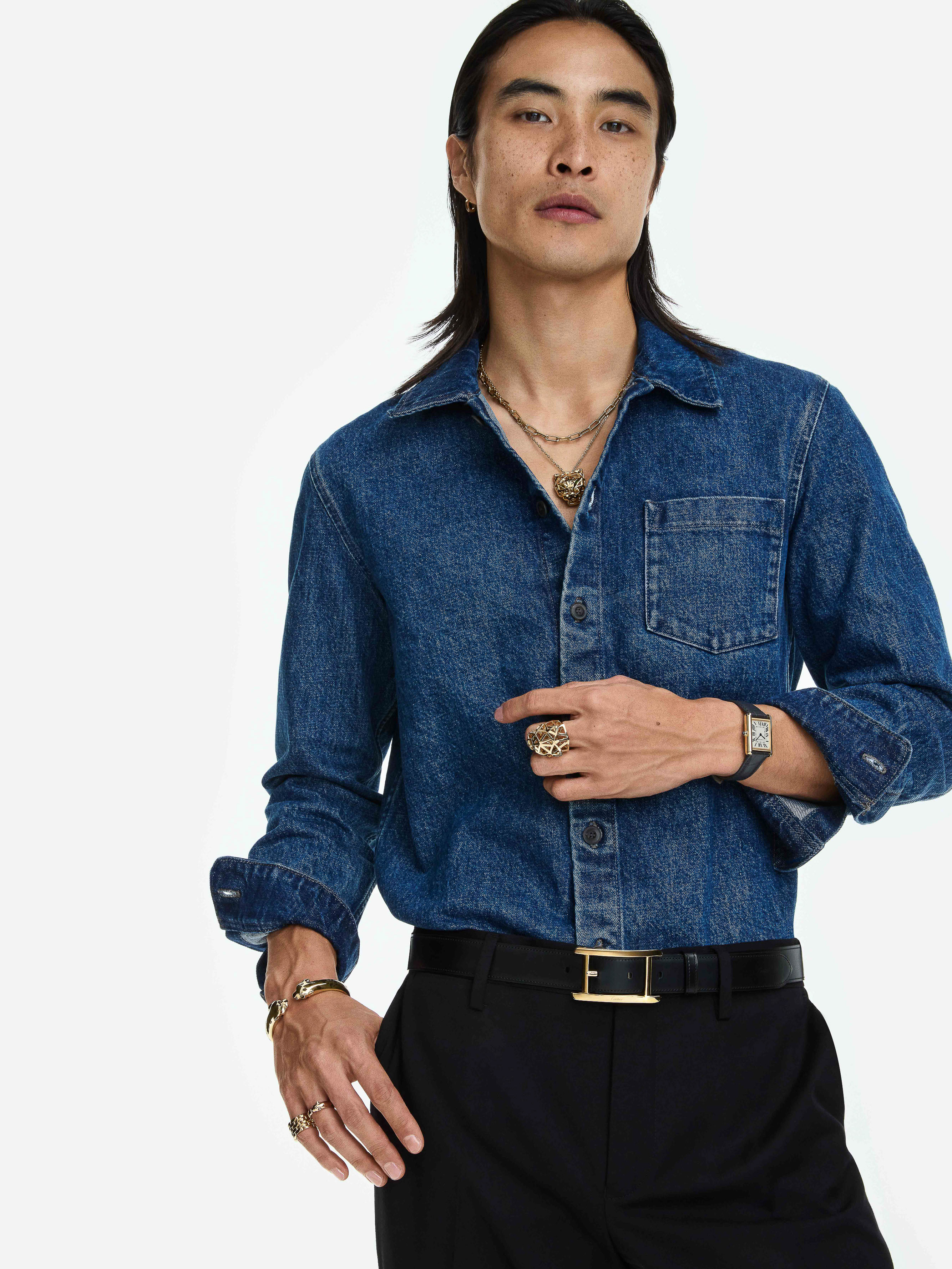 East Asian man in a blue denim shirt and black trousers wearing a gold necklace, watch, and rings against a white studio background.