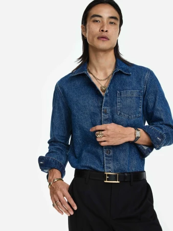 East Asian man in a blue denim shirt and black trousers wearing a gold necklace, watch, and rings against a white studio background.