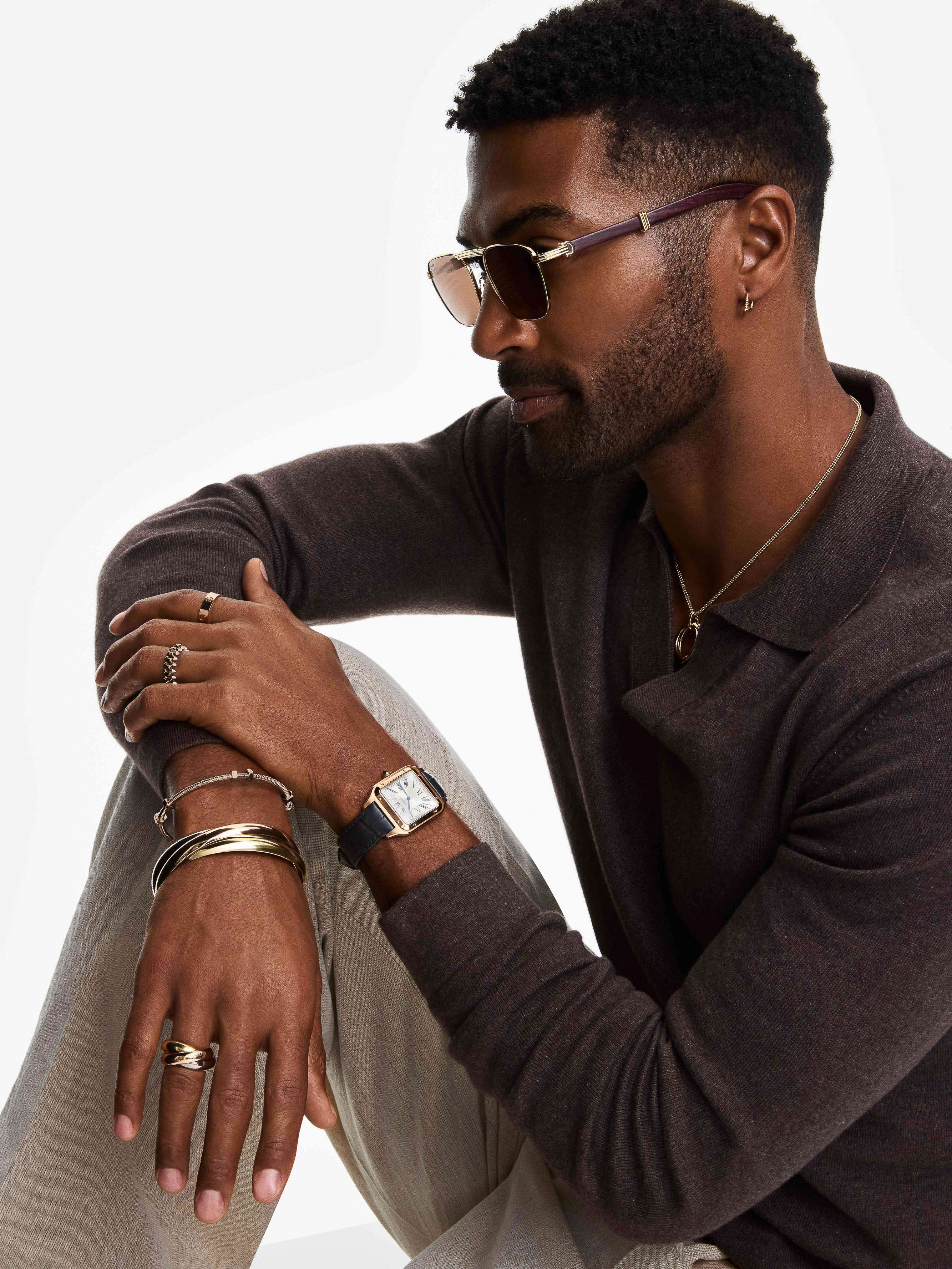 Stylish dark-skinned man with sunglasses, a brown collared shirt, jewelry, and a watch sits indoors in front of a white backdrop.