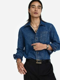 East Asian man in a blue denim shirt and black trousers wearing a gold necklace, watch, and rings against a white studio background.