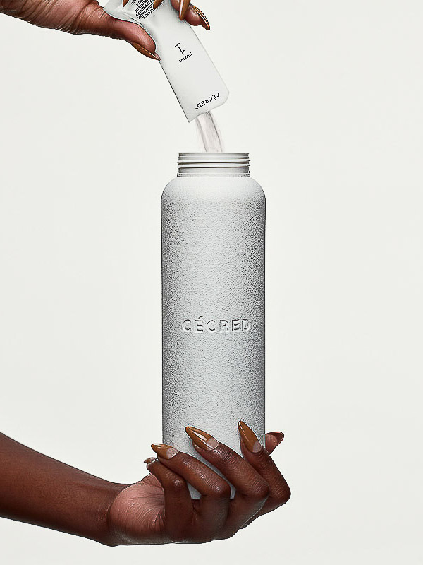 Close-up studio shot featuring hands pouring contents of a small white packet into a sleek, textured grey CÉCRED water bottle.