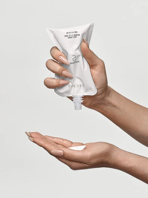Hands dispensing beauty treatment from a white tube onto the other hand, against a neutral background, showcasing personal care and beauty.