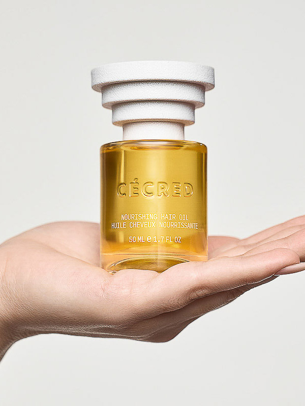 Close-up of a hand holding CÉCRED Nourishing Hair Oil bottle with a stacked cylindrical cap against a light gray background.