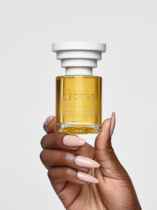 Close-up of dark-skinned hand holding clear bottle of Cécéd hair oil with white cap against a plain background in portrait orientation.