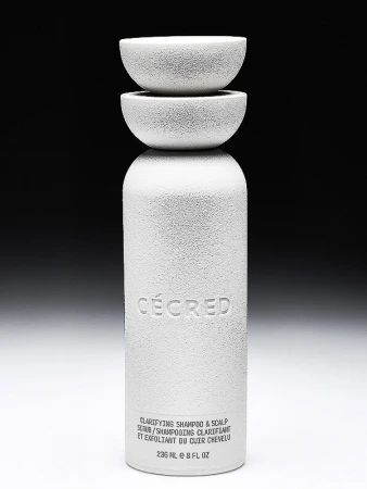 White bottle of clarifying shampoo and scalp scrub, topped with two small bowls, set against a gradient black to white background.