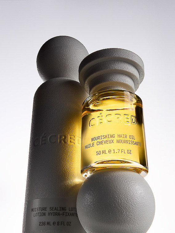 Close-up of CÉCRED moisturizing hair products: a small bottle of nourishing hair oil stacked on a white sphere, and a larger lotion.