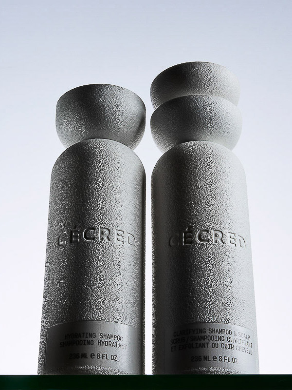 Two grey bottles of hair shampoo from Cécred brand with matching grey semi-spheres set against a bright white background.