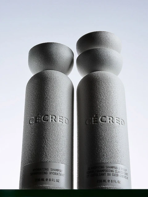 Two grey bottles of hair shampoo from Cécred brand with matching grey semi-spheres set against a bright white background.