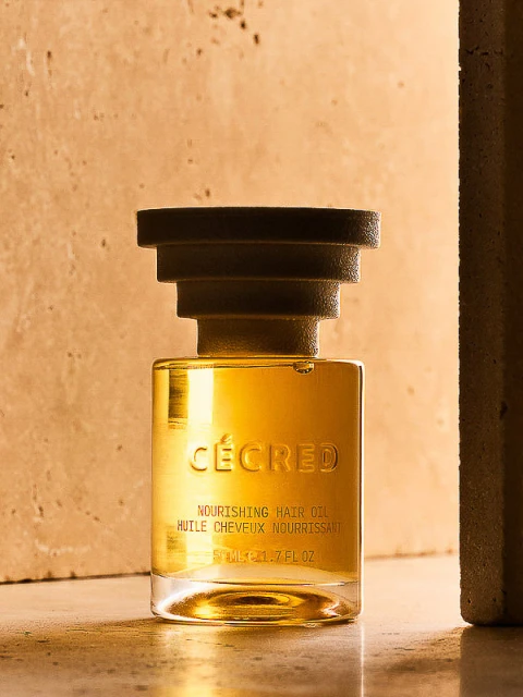 Elegant studio shot of Cécred Nourishing Hair Oil in a square glass bottle with a black cap, set against a neutral-toned textured backdrop.