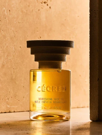 Elegant studio shot of Cécred Nourishing Hair Oil in a square glass bottle with a black cap, set against a neutral-toned textured backdrop.