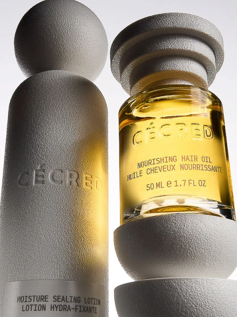 Two bottles of CÉCRED hair products, one featuring nourishing hair oil and the other moisture sealing lotion, isolated on a white backdrop.
