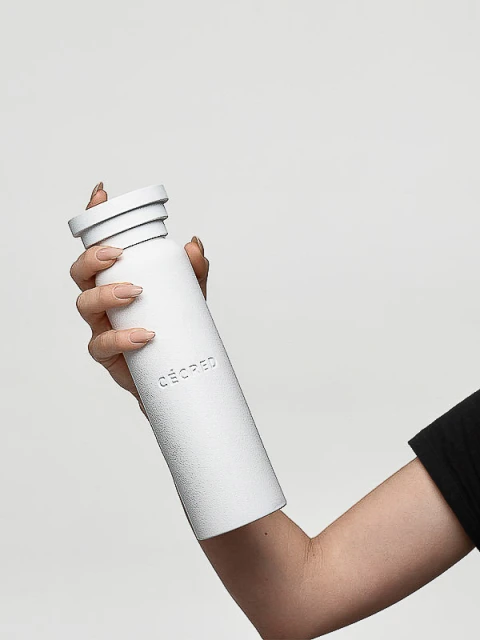 Woman holds textured white CÉCRED water bottle at eye-level against a white backdrop, showcasing minimalist product design and lifestyle.
