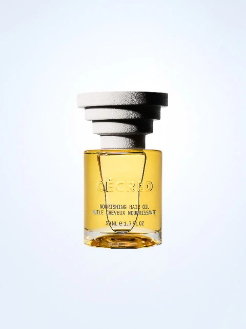 Close-up of a 'Décred' nourishing hair oil bottle with a layered white cap, showcasing its golden liquid and label against a light backdrop.