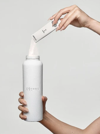 A woman's hands pour a white powder supplement from a packet into a white, textured metal water bottle against a solid grey backdrop.