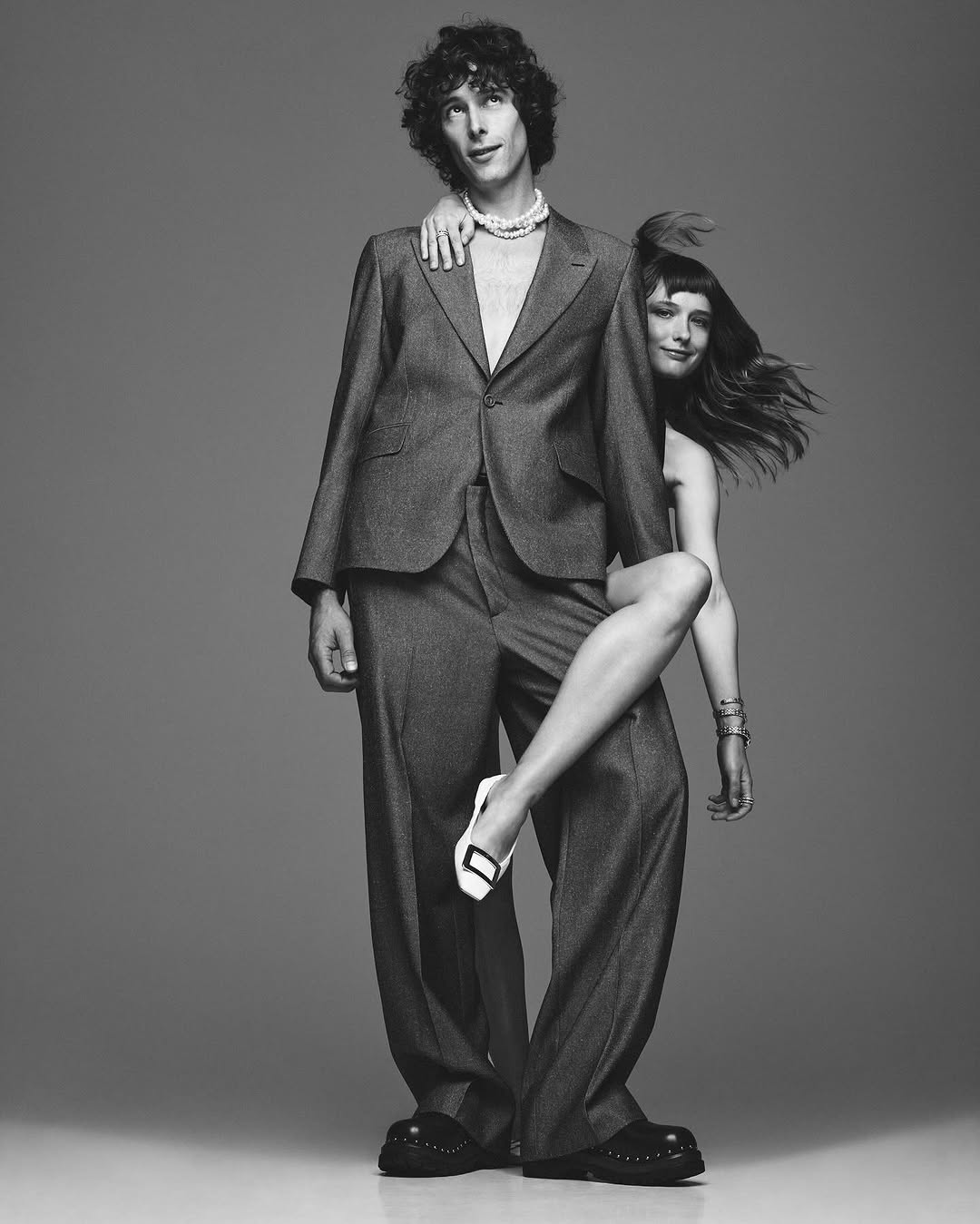 Monochrome studio shot of a tall man in a suit with a woman draped over his shoulder, both wearing pearl necklaces and staring ahead.