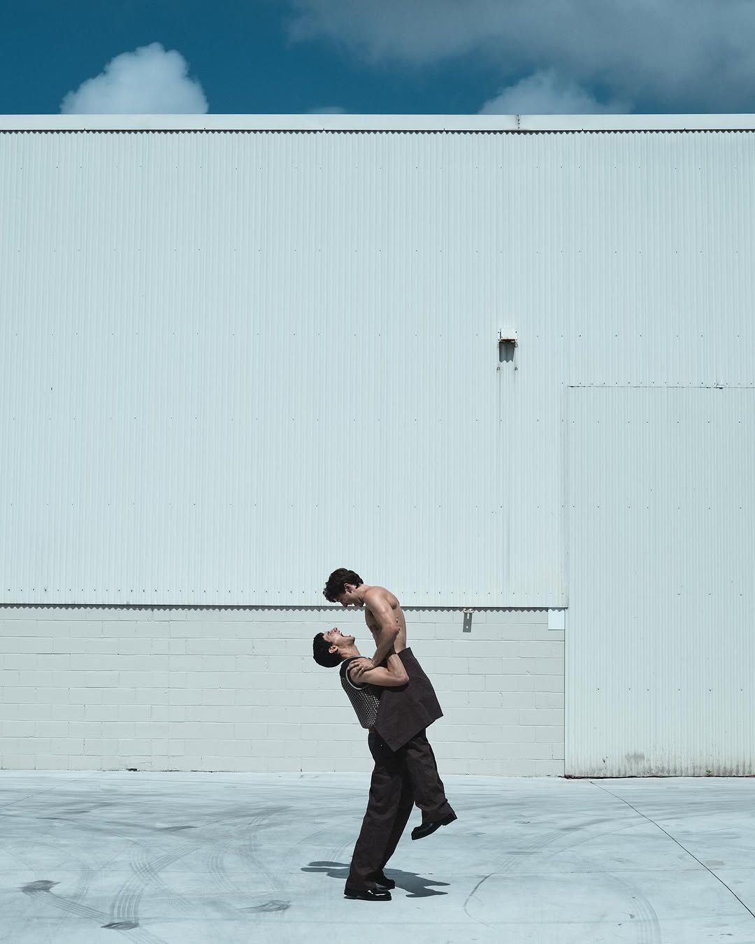 Two men are outdoors in daylight: one shirtless being held up by the other who is wearing a suit and shoes, against a white wall.