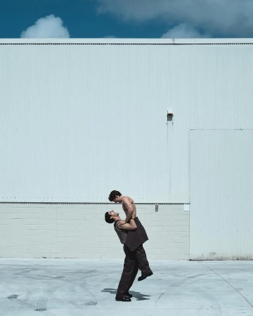 Two men are outdoors in daylight: one shirtless being held up by the other who is wearing a suit and shoes, against a white wall.