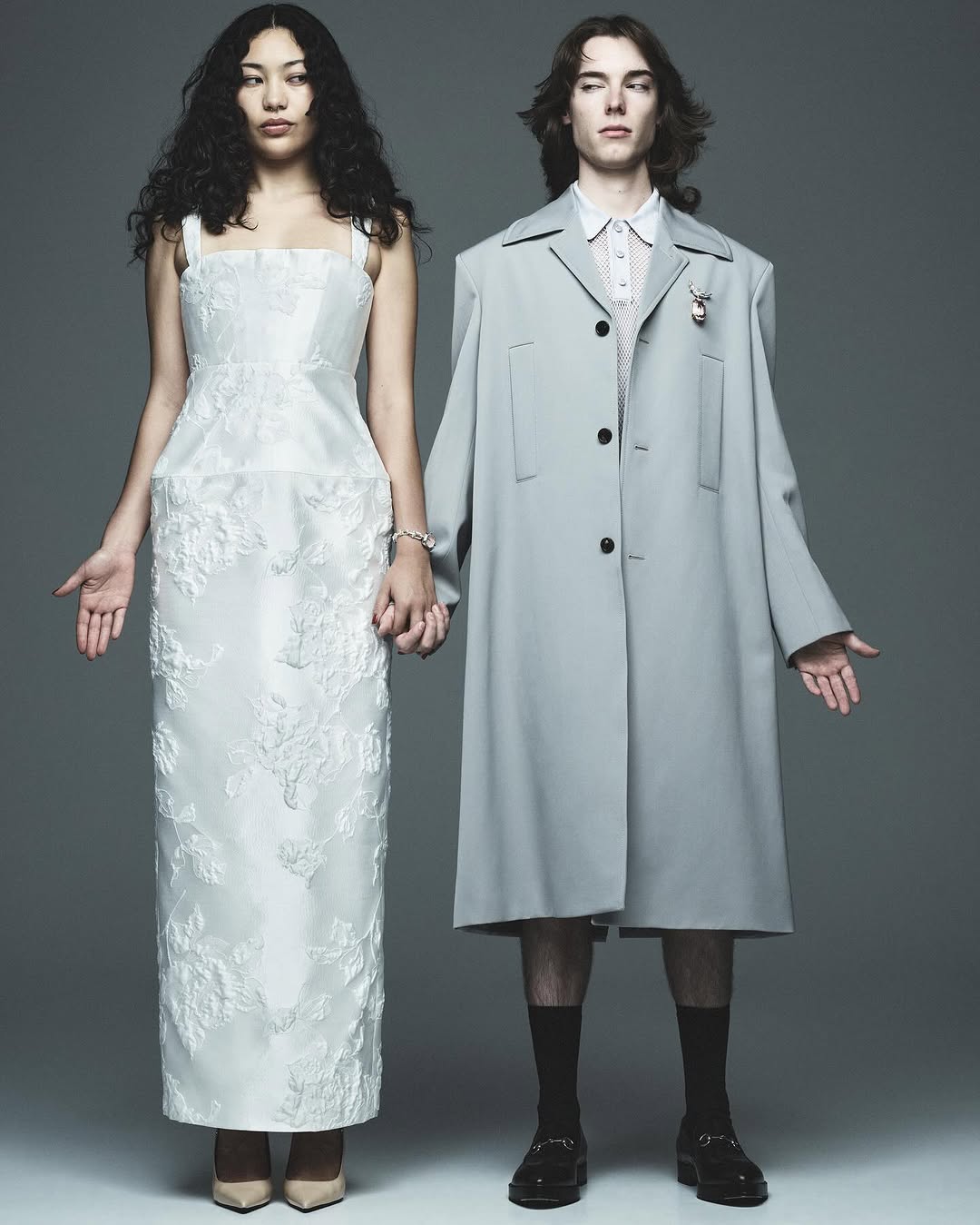 A couple poses in a photo studio. A woman wears a white gown with lace and the man is wearing a gray overcoat and loafers.