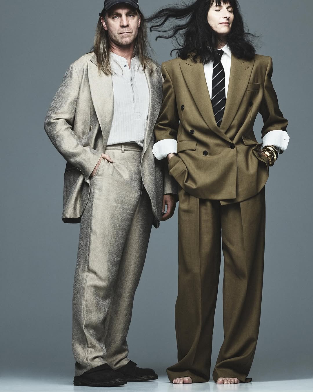Two people in suits stand in a studio shot. Man in a light suit and cap; woman in a brown suit and tie. Fashion editorial.