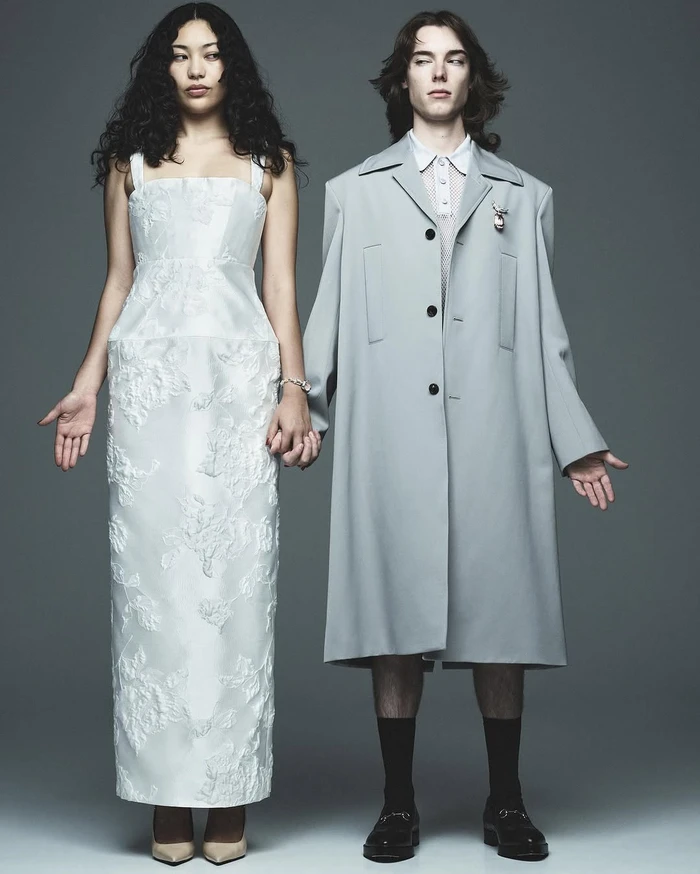 A woman in a white patterned dress and a man in a grey coat holding hands standing against a grey backdrop for Russh magazine.