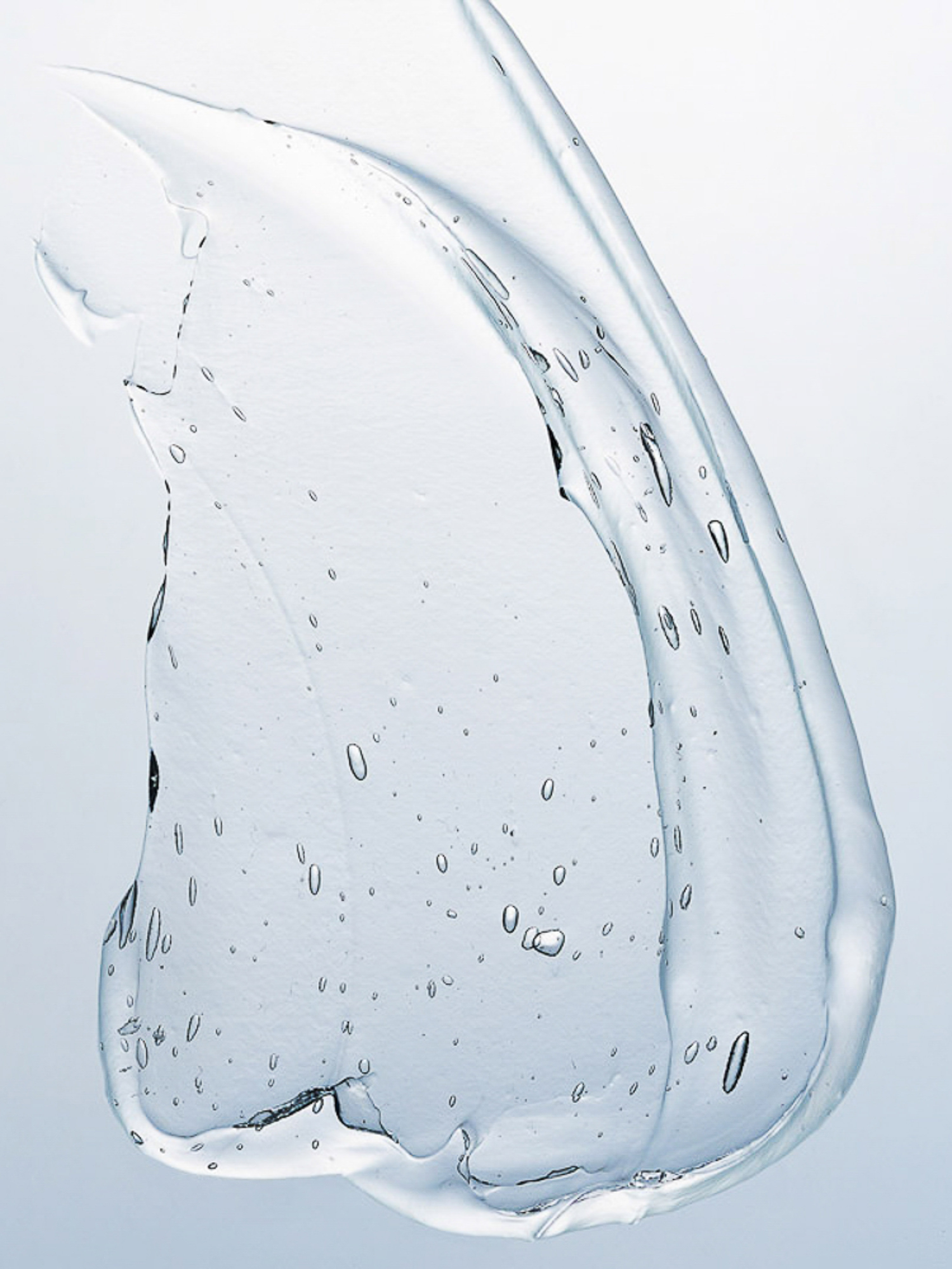Close-up of a clear, viscous gel smear against a light blue backdrop. The texture is transparent with tiny air bubbles visible throughout.