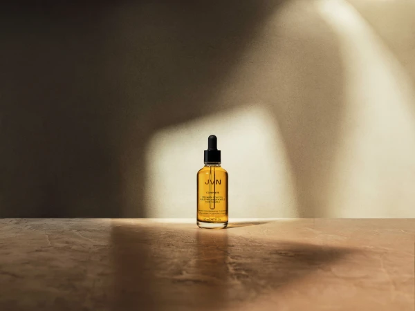 A bottle of JVN Complete Pre-Wash Scalp & Hair Treatment Oil on a reflective surface with soft lighting against a neutral background.