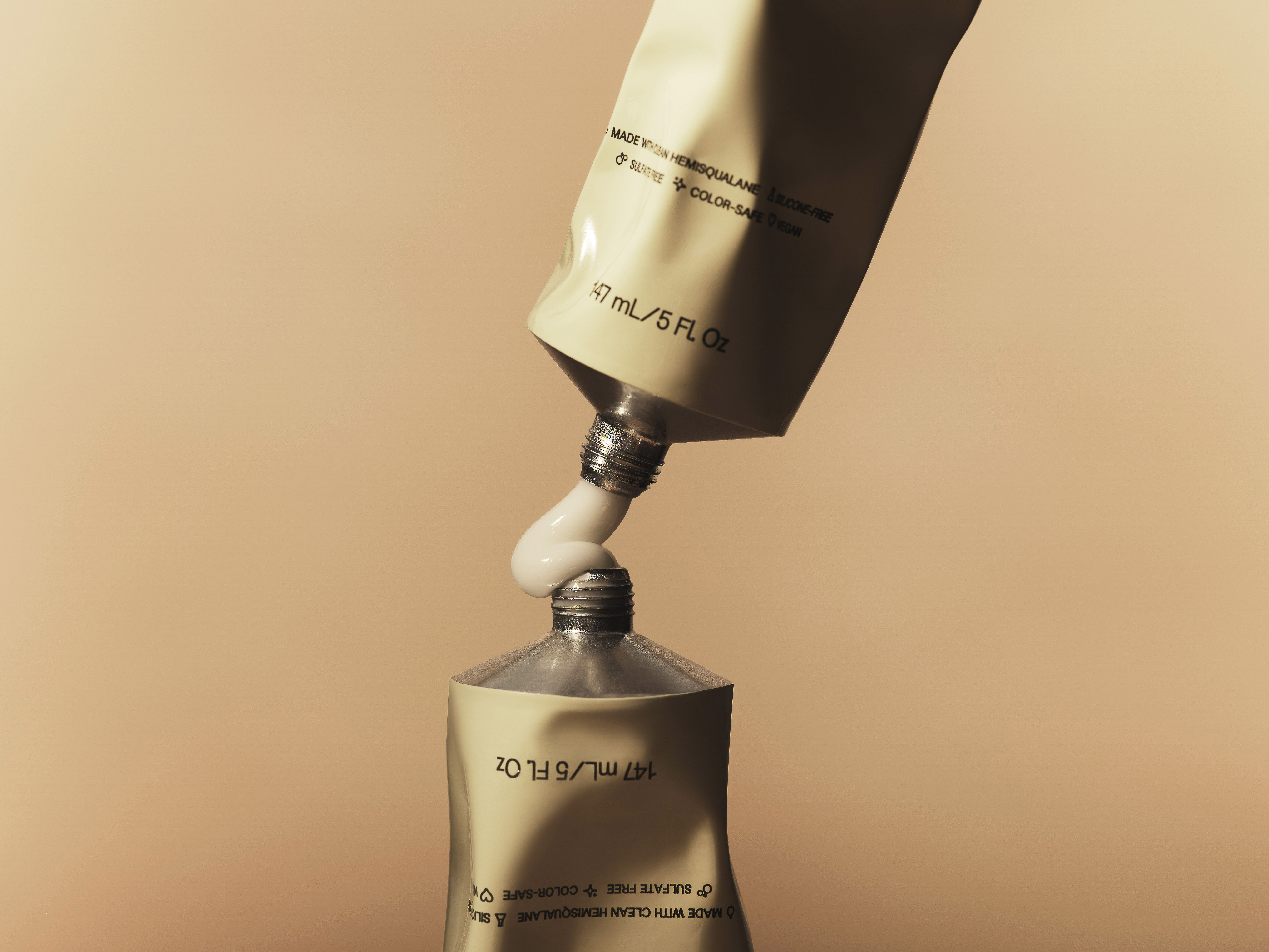 Two cosmetic cream tubes, one nearly inverted, releasing cream onto the other, close-up on a soft tan studio background, minimal.