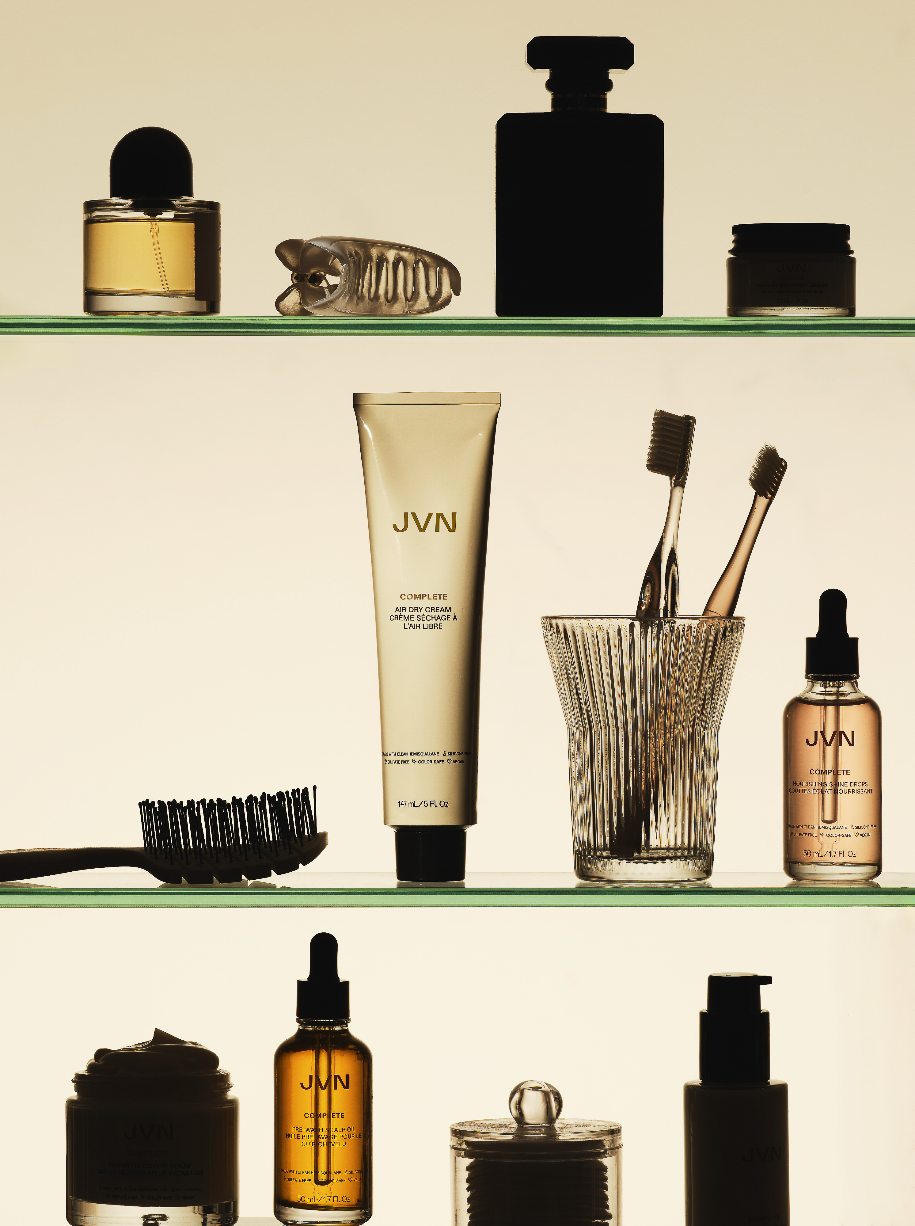 Still life arrangement of JVN brand haircare and skincare products on glass shelves including bottles, tubes, jars, brushes, and toothbrushes.